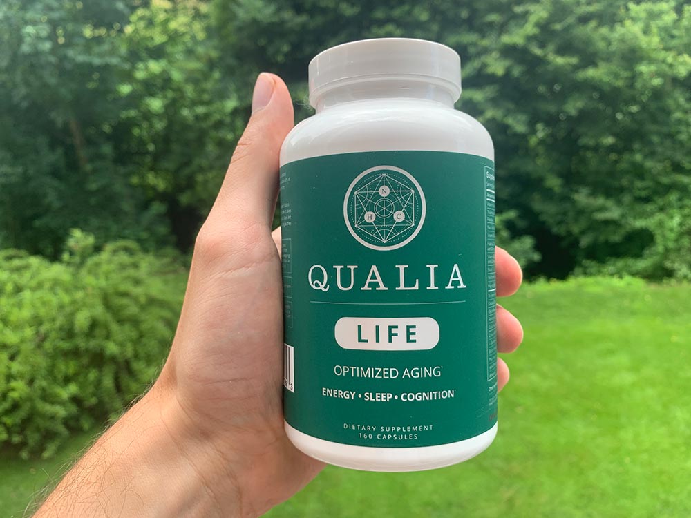 A hand holding one bottle of Neurohacker Qualia Life infront of a green backyard