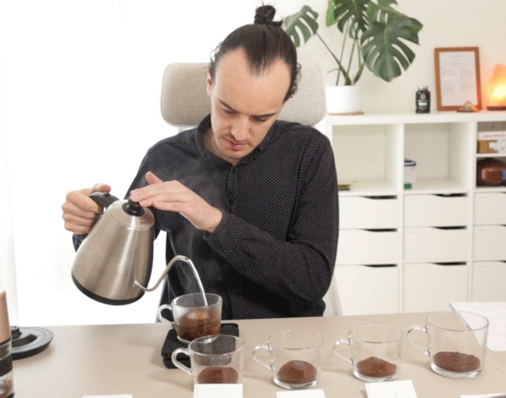 Max pouring hot water into a cup of ground coffee at a table with 5 cupping cups