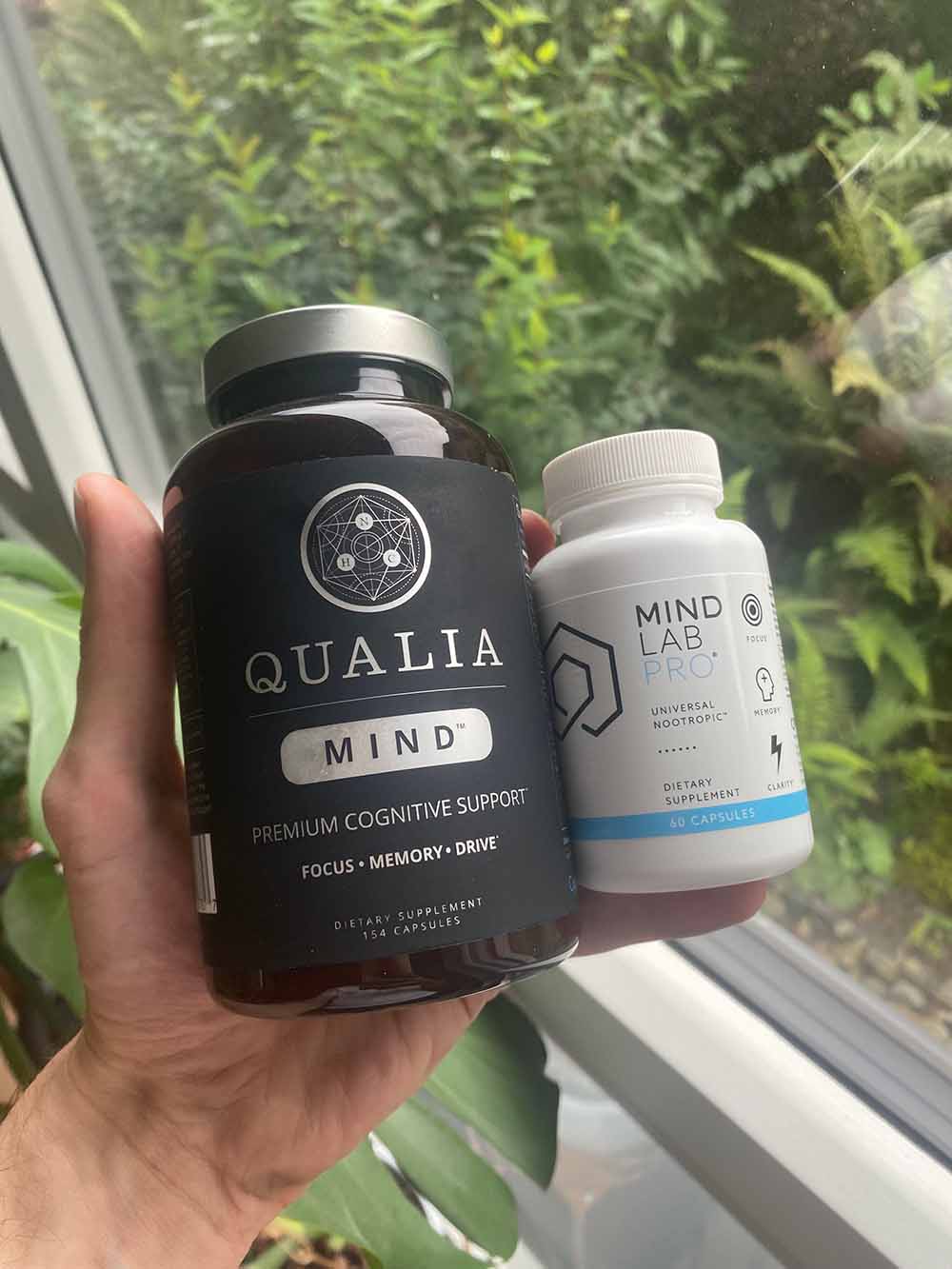 A hand holding a bottle of Qualia Mind and a bottle of Mind Lab Pro next to each other. Lush green background.