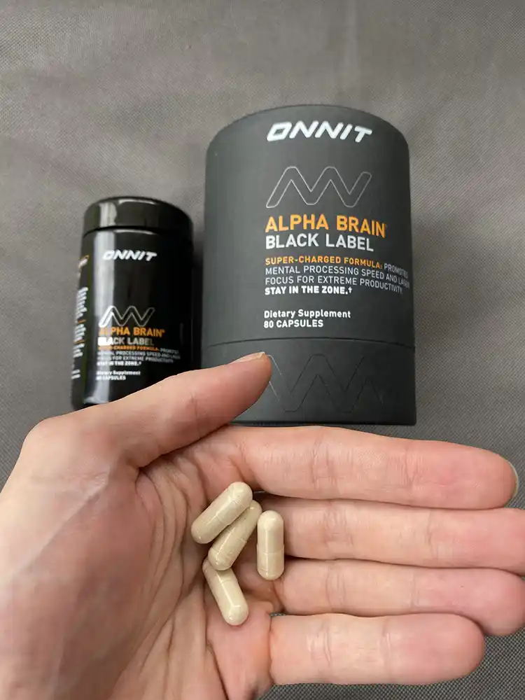 A hand holding 4 capsules of Alpha Brain Black Label with the Alpha Brain Black Label bottle and packaging in the background